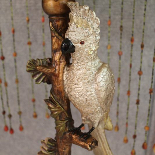 A table lamp with a decorative white cockatoo figurine perched on a brown branch with green leaf accents. Shade has hanging beaded strands.