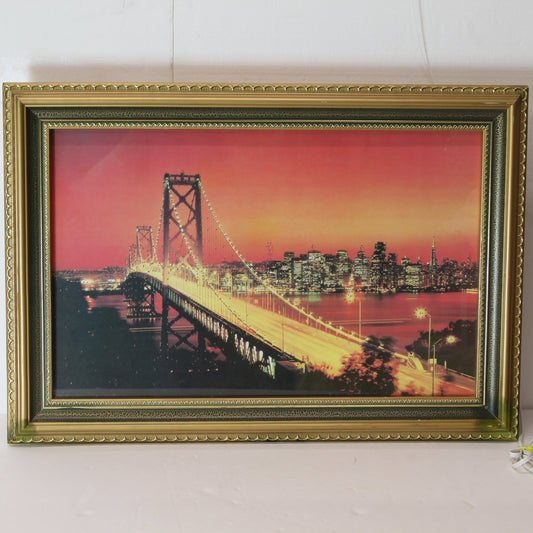 "Vintage 1980s San Francisco–Oakland Bay Bridge motion light box in an ornate gold frame. The image shows the bridge and city skyline at sunset with a pink and orange sky."