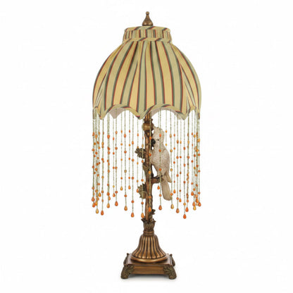 A decorative table lamp with a beige and olive green striped fabric shade, long strands of hanging beads, and a white bird figurine on an ornate bronze-toned base.