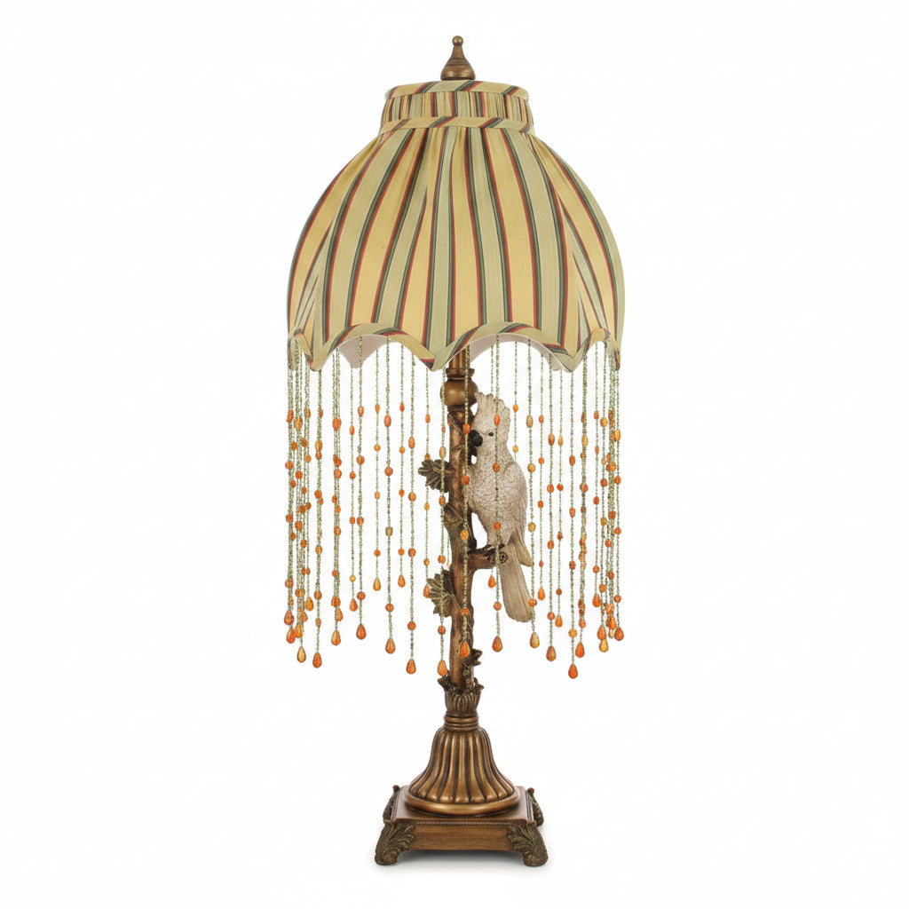 A decorative table lamp with a beige and olive green striped fabric shade, long strands of hanging beads, and a white bird figurine on an ornate bronze-toned base.