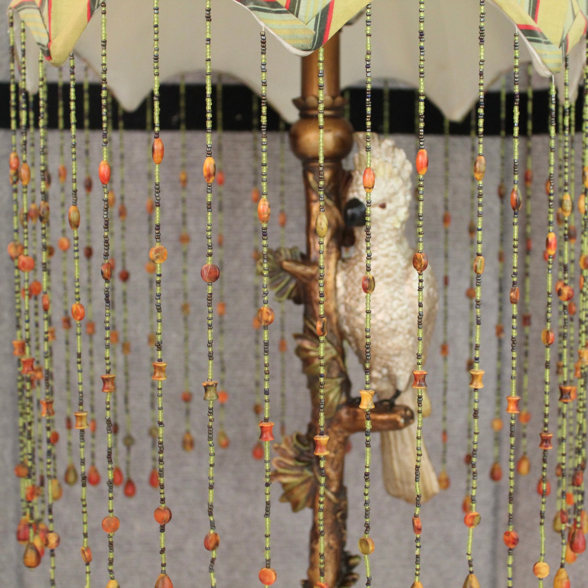 A close-up of a decorative table lamp with a white cockatoo parrot surrounded by vertical strands of beads in green, black, and orange. 