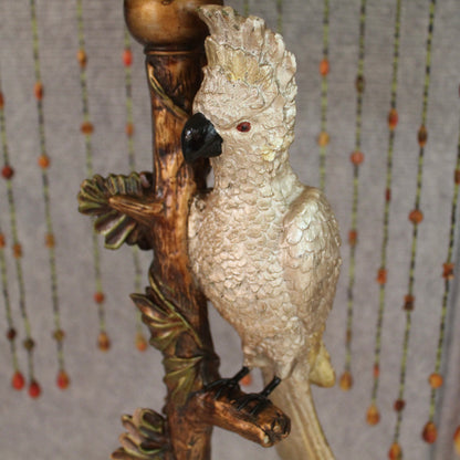 A table lamp with a decorative white cockatoo figurine perched on a brown branch with green leaf accents. Shade has hanging beaded strands.